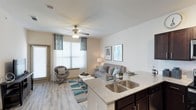 This single-floor residence features an open-concept layout that seamlessly connects the kitchen and living areas, creating a cohesive and functional environment. The kitchen is appointed with dark espresso cabinetry, a double stainless steel sink, and a granite-topped island that provides both additional workspace and a natural divide between the kitchen and living room.

The living area is finished with wood-style plank flooring that runs throughout the space, adding warmth and visual continuity. A ceiling fan with integrated lighting serves as a functional centerpiece overhead, while the textured ceiling adds subtle architectural detail. Large windows dressed with white blinds and teal accent curtains allow generous natural light to fill the room, contributing to the overall airy atmosphere.

The neutral color palette — composed of soft whites, warm beiges, and light grays — provides a versatile backdrop throughout. An exterior door in the living area suggests access to an outdoor space, enhancing both convenience and livability. Overall, the layout is designed to maximize the sense of openness while maintaining a warm, welcoming ambiance.