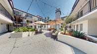 Located at 1318 N Mansfield Ave in the heart of Hollywood, Los Angeles, this two-story courtyard complex offers a generously sized open-air environment spanning a substantial ground-floor footprint. The property is organized around a series of interconnected outdoor courtyards, creating a welcoming, resort-like atmosphere flooded with Southern California sunlight.

The courtyards feature decorative stamped concrete paving in circular and geometric patterns, providing both visual interest and durable surface coverage throughout the common areas. Raised white planter beds are strategically placed across the space, filled with drought-tolerant and tropical plantings including agave, palms, cacti, and lush broadleaf greenery that lend a vibrant, landscaped character to the environment.

The crisp white stucco exterior walls frame the open courtyards, while dark-framed windows with louvered blinds line the ground-floor units. Bold navy blue iron railings accent the second-floor balconies, which overlook the courtyards below. Black metal staircases with wooden treads provide access between floors, adding an industrial-modern touch to the overall aesthetic.

The main gathering courtyard is furnished with patio seating and market umbrellas, with overhead string lighting that creates an inviting ambiance for evening use. A teal-painted elevator provides convenient vertical access. The combination of Mediterranean-inspired architecture, open-sky exposure, and thoughtful landscaping gives this Hollywood property a distinctive, community-centered character.