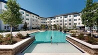 Located at 19101 Town Ridge Ln in Houston, TX, this outdoor amenity area is a standout feature of the modern multi-story residential community it serves. At its centerpiece is a generously sized resort-style swimming pool with crystal-clear blue water, framed by brick accent walls, entry steps, and a wide surrounding deck lined with lounge chairs — ideal for relaxing under the Texas sun. The pool is nestled within a curved architectural embrace of the building's contemporary exterior, finished in light gray board-and-batten siding with large windows that overlook the space. Adjacent to the pool, multiple distinct outdoor courtyard zones offer varied settings for recreation and leisure. Paver-lined plazas feature umbrella-shaded dining tables and seating, while open lawn areas with lush artificial turf provide space for lawn games such as cornhole. A covered pergola with wicker lounge furniture offers a shaded retreat, and string lights overhead create an inviting ambiance for evening gatherings. Thoughtfully curated landscaping throughout — including ornamental trees, magnolias, shrubs, and layered plantings — adds natural color and texture to the environment, creating a polished, resort-like atmosphere in the heart of the Houston, TX community.