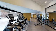 This fitness center offers a comprehensive workout environment designed with both functionality and aesthetics in mind. The space features a single open floor plan with a clean, modern design highlighted by a drop-tile ceiling with recessed lighting and a large floor-to-ceiling window along one wall, allowing natural light to fill the room and providing views of the surrounding outdoor scenery.

A row of cardio machines, including treadmills, ellipticals, and stationary bikes, lines the windowed wall, allowing users to exercise while enjoying the outside view. The flooring consists of durable rubber gym matting, practical for both equipment placement and user safety.

Towards the interior of the space, a well-organized free weight section includes a full dumbbell rack alongside adjustable benches, catering to strength training needs. Warm wood-paneled accent walls add a refined visual contrast to the otherwise neutral tones of the space.

Additional strength and cable machines are visible toward the back, making use of mirrored walls to enhance the sense of depth and space. Overall, the fitness center presents a clean, well-maintained, and thoughtfully organized environment suitable for a full range of workout routines.
