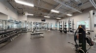 Located at 25 Woodridge Crescent in Ottawa, Ontario, this well-equipped fitness facility offers a generous, open-plan training floor designed to accommodate a wide variety of workout styles. The space features soaring exposed ceilings with industrial-style steel trusses, creating an airy and expansive atmosphere throughout. Bright fluorescent lighting is suspended from the ceiling grid, ensuring the entire floor is evenly and thoroughly illuminated.

The facility is outfitted with a robust selection of strength training equipment, including multiple power racks loaded with weight plates, barbells, cable machines, and a full dumbbell rack with adjustable benches. Functional training stations are also present, featuring suspension trainers, stability balls, and abdominal equipment. Wall-mounted flat-screen televisions are positioned throughout the space for convenience.

Full-length mirrors line key sections of the walls, enhancing the sense of openness while allowing users to monitor their form during training. The floor is finished with durable rubber sport flooring, providing both safety and performance underfoot. Windows along the perimeter admit natural light and offer views of the surrounding greenery. Industrial wall-mounted fans contribute to ventilation and comfort. The overall ambiance is clean, professional, and purpose-built for serious fitness training.