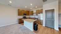 This single-floor unit offers a well-designed, open layout that connects the kitchen and living areas seamlessly. The kitchen is a standout feature, showcasing warm medium-toned flat-front cabinetry with sleek hardware, granite countertops with subtle veining, and a full suite of stainless steel appliances including a range, over-the-range microwave, dishwasher, and refrigerator. A center island with a matching granite surface provides additional prep space and casual seating potential. Light wood-look flooring runs throughout the kitchen, transitioning to plush carpet in the living and bedroom areas.

The living area is generously sized, bathed in natural light from multiple windows fitted with horizontal blinds that offer views of the surrounding neighborhood. A textured ceiling with recessed lighting adds to the bright, clean aesthetic throughout the space.

The bedroom features soft carpeting, a ceiling fan, and a window providing additional natural light and exterior views. The bathroom includes a white tub and shower surround, a toilet, granite-topped vanity, and brushed nickel fixtures and grab bar, offering both style and accessibility. Overall, the unit presents a clean, contemporary atmosphere with consistent neutral tones throughout.
