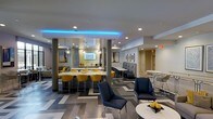 Located on 9th Street in Durham, NC, this single-floor space offers a thoughtfully designed interior that blends contemporary aesthetics with functional versatility. The open-plan layout creates a welcoming communal environment, highlighted by a bold chevron-patterned floor in shades of gray that anchors the entire space with a dynamic visual element.

A central bar or counter area serves as a focal point, lined with striking yellow upholstered barstools that provide a vivid contrast against the neutral surroundings. Above, pendant globe lights hang at varying heights, complementing the recessed ceiling lighting throughout. A standout architectural detail is the vibrant blue LED cove lighting that traces the perimeter of a dropped ceiling section, lending the space a modern, energetic ambiance.

Seating is arranged in distinct zones to accommodate a variety of uses, from casual lounging to collaborative work or dining. Navy blue accent chairs, banquette-style seating, and high-top tables with stools are thoughtfully distributed across the floor. Decorative wall art and patterned wallcoverings add personality and depth to the interior, while structural columns are seamlessly integrated into the design. The overall atmosphere is polished, social, and contemporary.