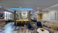 Located on 9th Street in Durham, NC, this single-floor space offers a thoughtfully designed interior that blends contemporary aesthetics with functional versatility. The open-plan layout creates a welcoming communal environment, highlighted by a bold chevron-patterned floor in shades of gray that anchors the entire space with a dynamic visual element.

A central bar or counter area serves as a focal point, lined with striking yellow upholstered barstools that provide a vivid contrast against the neutral surroundings. Above, pendant globe lights hang at varying heights, complementing the recessed ceiling lighting throughout. A standout architectural detail is the vibrant blue LED cove lighting that traces the perimeter of a dropped ceiling section, lending the space a modern, energetic ambiance.

Seating is arranged in distinct zones to accommodate a variety of uses, from casual lounging to collaborative work or dining. Navy blue accent chairs, banquette-style seating, and high-top tables with stools are thoughtfully distributed across the floor. Decorative wall art and patterned wallcoverings add personality and depth to the interior, while structural columns are seamlessly integrated into the design. The overall atmosphere is polished, social, and contemporary.