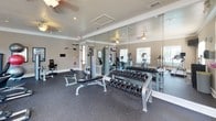 This single-floor fitness center is thoughtfully designed to support a comprehensive workout experience. The main exercise area is anchored by a full rack of dumbbells paired with an adjustable weight bench, ideal for strength training, alongside a multi-function cable machine for a wide range of resistance exercises. Large floor-to-ceiling mirrors line the walls, visually expanding the space and allowing users to monitor their form during workouts. The rubber-floored cardio zone is outfitted with multiple True-brand treadmills and elliptical trainers positioned near windows with blinds, providing natural light and outside views during exercise. Ceiling fans throughout the space help maintain a comfortable environment. A dedicated functional training area includes a stationary recumbent bike, a rack of medicine balls in various weights, stability balls, yoga mats, and wall-mounted exercise charts — all positioned beneath a wall-mounted television for entertainment. The facility features warm neutral-toned walls, crown molding, and a clean, well-maintained aesthetic. A wood-framed glass entry door provides access, and safety features such as a fire extinguisher and emergency exit signage are present throughout.