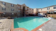 This residential community is defined by its inviting outdoor amenity space, centered around a large, sparkling swimming pool with clear turquoise water. The pool area is framed by a classic brick coping surround and enclosed within a secure iron perimeter fence, lending the space both safety and a polished aesthetic. A shaded seating area with a colorful umbrella provides a comfortable spot for relaxation poolside.

The surrounding multi-story buildings feature a warm combination of tan siding and brick detailing, giving the exterior a traditional yet welcoming character. Each level is equipped with private balconies framed by iron railings, offering residents outdoor space and views of the courtyard and pool below. The architectural design reflects a cohesive and well-maintained community aesthetic throughout.

The grounds are neatly landscaped with greenery accenting the base of the buildings, contributing to an overall sense of order and care. The open layout of the outdoor area allows for ample natural light throughout the day, enhancing the bright and airy atmosphere of the community. Whether relaxing by the pool or enjoying the fresh air from a private balcony, this community offers a range of outdoor living experiences well-suited for comfortable, everyday residential life.