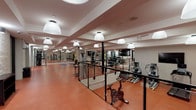 This fitness center offers a comprehensive workout environment spread across a single floor. The space is defined by its warm terracotta rubber flooring, which provides both durability and comfort underfoot throughout the facility. Structural columns and exposed ceiling beams add an industrial character, while large pendant dome lights and recessed fixtures ensure the area is well-illuminated.

The main workout floor is generously proportioned and houses a diverse range of equipment, including treadmills, elliptical trainers, and a stationary cycling bike. A wall-mounted flat-screen television is positioned to face the cardio equipment, enhancing the workout experience. Full-length mirrors line one wall, visually expanding the space and allowing users to monitor their form during exercise.

A dedicated strength training zone features a cable machine system, weight rack with dumbbells, and a bench press setup. A separate enclosed cycling studio, delineated by glass partitions and black metal framing, provides a quieter space for focused cycling sessions. Painted white brick walls add texture throughout, complementing the neutral wall tones. Climate control is addressed through wall-mounted split air conditioning units, and the space is accessible via a glass-paneled entry door.