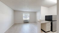 Located at 703 N Shore Dr in Jeffersonville, Indiana, this apartment offers an efficient, single-floor layout comprising a living room, kitchen, hallway, bathroom, and closet. The living area is bright and open, featuring light wood-look flooring, neutral white walls, and a textured ceiling. A window fitted with horizontal blinds allows natural light to filter in and offers a view of the surrounding greenery, while a wall-mounted HVAC unit provides convenient climate control directly beneath it. The open layout allows for flexible furniture arrangement, making the most of the available footprint. The kitchen is positioned adjacent to the living area and features granite-style countertops with a breakfast bar that opens toward the main living space, creating a connected, functional flow. The bathroom is accessible via the hallway and includes a full bathtub and shower combination equipped with a safety grab bar, a toilet, and a vanity with a granite-style countertop, all finished with matching light flooring. A dedicated closet provides organized storage within the unit. Throughout the apartment, the consistent light flooring, clean white walls, and neutral palette create a cohesive, understated aesthetic that is easy to personalize.