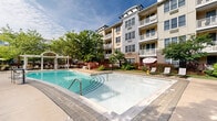 Located at 50 Chelsea Ave in Long Branch, New Jersey, this outdoor amenity area serves as a central gathering space within a multi-story residential community. The centerpiece is a generously sized swimming pool with crystal-clear blue water, accessible via wide entry steps with stainless steel handrails. The pool deck is finished in a combination of paved stone and concrete, providing ample space for lounging and socializing. Rows of chaise lounges with accent tables are arranged along the poolside, offering residents comfortable spots to relax and enjoy the open sky. A charming pergola structure anchors one corner of the deck, adding architectural interest and a shaded retreat. The area is enclosed by a distinctive red metal fence lined with vibrant seasonal plantings, including flowering perennials that add color and life to the space. Mature trees are thoughtfully positioned throughout, offering natural shade and a sense of privacy. The surrounding multi-story building features balconies that overlook the pool courtyard, creating an intimate, resort-like atmosphere. Landscaped greenery softens the hardscape and enhances the overall ambiance, making this outdoor space a well-designed and inviting community amenity.