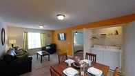 Located at 3800 Bostic Dr in Greenville, NC, this single-floor residence offers a well-organized and functional layout designed for comfortable everyday living. The open-concept living and dining area serves as the heart of the space, featuring warm accent walls in a bold orange hue that add energy and character to the interior. The dining area accommodates a full-sized table with seating for multiple guests, while the adjacent living room provides a cozy retreat with carpeted flooring, a wall-mounted flat-screen television, and a ceiling-mounted light fixture. Natural light filters in through a window dressed with green curtains, brightening the shared living space. A bedroom is accessible directly from the main living area, offering a ceiling fan for added comfort. One of the standout functional features of this unit is the in-unit laundry area, which includes a full-sized washer and dryer along with shelving for storage. The textured ceilings and consistent flooring throughout contribute to a cohesive and tidy aesthetic. Overall, this space balances practicality with a welcoming atmosphere, making it a solid residential option in Greenville, NC.