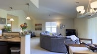This single-floor residence offers a well-designed open-concept layout that promotes a sense of spaciousness and easy flow between living areas. The living room is a central highlight, featuring vaulted ceilings that add vertical dimension and an airy feel to the space. Soft, neutral carpet flooring runs throughout, contributing to a warm and comfortable atmosphere. A large sectional sofa anchors the living area, which is brightened by a window that allows natural light to fill the room. A flat-screen television is positioned for comfortable viewing, and wall art adds a decorative touch to the neutral-toned walls. A pendant light fixture hangs gracefully, complementing the room's lighting scheme. Adjacent to the living area, a partial wall with a granite countertop separates the kitchen, which features dark stone surfaces that contrast with the lighter tones of the surrounding space. A dining area is also nearby, completing the open-plan arrangement. Overall, the home presents a cohesive and functional living environment with a relaxed, inviting ambiance suited for everyday living.