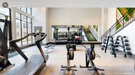 Located at 330 Towerview Ct in Cary, NC, this two-story fitness center offers a generously sized, contemporary workout facility designed for a comprehensive exercise experience. The ground floor welcomes visitors with soaring ceilings, large floor-to-ceiling windows that flood the space with natural light, and views of the surrounding community. A striking botanical wall mural flanks an open staircase, adding an artistic focal point to the space. The lower level is equipped with rowing machines, free weights, benches, cable machines, and dedicated functional training zones with battle ropes and plyo boxes. A full-length mirror wall enhances the sense of openness in the weight training area. The upper floor continues the modern aesthetic, featuring distinctive oval ring lighting fixtures suspended from the ceiling, creating an ambient and energizing atmosphere. This level houses additional cardio equipment, including treadmills, air bikes, and cable machines, alongside a strength training section with squat racks and barbells. Large windows on the upper floor offer scenic outdoor views. The facility also includes a well-appointed restroom with herringbone-patterned tile walls, gold-accented fixtures, and elegant pendant lighting, reflecting the same refined design sensibility found throughout the gym.