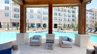 This outdoor amenity space is designed to deliver a resort-like experience, combining multiple recreational zones into a cohesive and visually impressive environment. The centerpiece is a generously sized swimming pool with vivid blue water, surrounded by wide open decking that accommodates rows of chaise lounges and shaded umbrella seating. A second pool area features in-water sun loungers positioned along a tiled shallow shelf, adding a leisure-forward design element. A freestanding gazebo with a tile roof and warm wood detailing serves as the focal point between the two pools, providing a shaded gathering space furnished with comfortable seating. An additional covered cabana with exposed timber beams and stone columns overlooks the main pool, offering wicker lounge chairs and a central accent table. Adjacent to the pool deck, a dedicated spa or fire feature area is framed by rocking chairs, lounge seating, and patio dining. A separate landscaped lawn zone features hammocks suspended beneath terracotta sail shades strung with bistro lights, creating a relaxed retreat with lush greenery and garden plantings throughout.