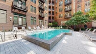 Located at 611 S Charles St in Baltimore's vibrant South Baltimore neighborhood, this outdoor amenity space serves as a central courtyard retreat enclosed by a multi-story brick residential building. The centerpiece is a generously sized rectangular swimming pool with crystal-clear blue water, framed by a clean stone coping surround and elevated pool deck. A dedicated sun deck lines one side of the pool, furnished with rows of modern white lounge chairs and chaise lounges, providing ample space for relaxation and sunbathing. Mature trees and well-maintained greenery are integrated throughout the courtyard, softening the urban setting and offering natural shade. An outdoor dining and grilling area is situated along the building's perimeter, featuring long bar-style countertops, multiple seating arrangements, and direct access through large glass-paneled doors — a practical space ideal for social gatherings. Warm brick façades with large windows and private balconies rise on all sides, creating a sheltered, courtyard-style environment with a strong sense of community. The Bocce Courtyard, visible via exterior signage, adds a recreational element to this well-appointed outdoor space. Paved stone walkways connect all areas seamlessly, making this a versatile and inviting outdoor amenity for residents.