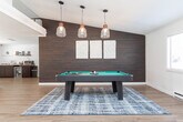 Located at 301 W Vandalia Rd in Greensboro, NC, this single-floor community space offers a well-organized and thoughtfully designed layout ideal for gathering and recreation. The main living area serves as the heart of the space, featuring a white-mantled fireplace with a wall-mounted television, plush velvet seating, and a patterned area rug over light hardwood-style flooring. Crown molding and clean white walls lend a polished, cohesive aesthetic throughout. Natural light flows in through multiple windows and glass-paneled doors that open to the outdoors, enhancing the open and airy feel of the lounge. Adjacent to the living area is a dedicated recreation room distinguished by a striking dark wood accent wall and pendant lighting, which frames a full-size pool table — a standout feature of the space. A workstation area is also present within this room. The kitchen is equipped with dark cabinetry, granite-style countertops, a beverage refrigerator, and pendant lighting, providing ample prep and serving space. The dining room near the entry serves as a welcoming reception area, complete with a glass-top table, mirrored wall panels, and a pendant light fixture. Hardwood-style flooring runs consistently throughout, unifying the space with a warm, contemporary character.