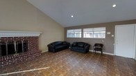 This single-story space centers around a spacious living area defined by striking architectural details and functional design. The room features vaulted ceilings that create an open, airy atmosphere, drawing the eye upward and adding a sense of generous volume to the interior.

A prominent brick fireplace anchors one side of the room, serving as a classic focal point with its full brick surround and white-painted mantle. The warmth of the brick contrasts tastefully with the neutral, beige-toned walls that run throughout the space.

The flooring consists of earthy, terracotta-toned tiles laid in a varied pattern, adding texture and visual interest underfoot while contributing to the room's warm overall palette. Recessed lighting in the ceiling supplements the natural light that filters in through a pair of wide windows along the far wall, offering views to the exterior and keeping the space bright throughout the day.

The layout provides ample room for seating arrangements, as evidenced by the placement of two sofas and an accent chair. The combination of the vaulted ceiling, fireplace, tiled floors, and natural light gives the space a comfortable, inviting character suited for everyday living.