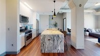 Located at 292 W Western Ave in Muskegon, Michigan, this contemporary urban unit offers a thoughtfully designed open-concept layout highlighted by rich hardwood floors and soaring ceilings throughout. The kitchen is a true centerpiece, featuring a dramatic full-slab granite waterfall island, sleek white cabinetry, stainless steel appliances, a mosaic tile backsplash, and striking black pendant lighting that adds an industrial-modern touch. Exposed ductwork and structural columns pay homage to the building's urban character.

The spacious living area is bathed in natural light from oversized floor-to-ceiling windows that frame sweeping views of the Muskegon cityscape and waterfront beyond. The space accommodates generous seating and a flat-screen television, making it ideal for both relaxing and entertaining.

Two well-appointed bedrooms offer a calm retreat, each furnished with comfortable queen beds, warm ambient lighting, and curated artwork. One bedroom enjoys direct access to the panoramic city views through its large window wall. Both bathrooms are finished with granite-topped white vanities, hardwood flooring, and clean, modern fixtures. The overall ambiance balances industrial loft aesthetics with warm, residential comfort throughout.