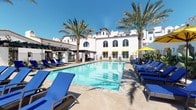 This amenity facility showcases a thoughtfully designed resort-style environment centered around a large, pristine outdoor swimming pool. The pool area is framed by tall palm trees and complemented by an expansive sun deck lined with blue lounge chairs and yellow-accented umbrellas, all set against a striking white Spanish Colonial-style building featuring arched windows, terracotta roof tiles, and covered pergola spaces for shade. The cheerful, sun-soaked atmosphere is enhanced by the vivid contrast of blue water, bright loungers, and lush greenery under clear skies.

The fitness center is a standout feature, offering a comprehensive, well-equipped workout space spread across a generously sized floor plan. Rows of cardio machines — including StairMaster climbers, ellipticals, and treadmills from True Fitness — line the perimeter near arched windows that allow ample natural light to fill the space. Floor-to-ceiling mirrors amplify the sense of openness throughout. A dedicated strength-training zone houses a full array of cable machines and resistance equipment, catering to a wide range of workout routines. Rubber flooring, recessed lighting, and a clean, modern finish complete the professional-grade fitness environment.