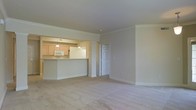 This single-floor residence offers a thoughtfully designed open-concept layout that seamlessly connects the living and kitchen areas. The spacious living room is finished with plush wall-to-wall carpeting in a warm neutral tone, creating a comfortable and inviting atmosphere. Crown molding runs along the ceiling, adding a refined architectural detail to the space.

The kitchen is positioned adjacent to the living area and features light wood cabinetry, a built-in microwave, and a full-sized refrigerator. Pendant lighting hangs above the kitchen bar, providing both functional illumination and a decorative touch. The bar-style counter offers a natural transition point between the kitchen and the living space, making it ideal for casual dining or entertaining.

The interior color palette consists of soft, neutral whites and beiges throughout, giving the space a clean and cohesive look. The raised panel doors and white trim complement the overall design aesthetic. The layout includes interior doors that suggest additional rooms beyond the main living area, allowing for flexible use of the space. Overall, the residence combines practical design with comfortable finishes, creating a welcoming and functional living environment.