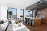 This single-floor high-rise residence is defined by its striking modern design and exceptional panoramic views, including city skylines, neighboring skyscrapers, and distant water vistas. Floor-to-ceiling windows fitted with navy-blue frames run throughout the unit, flooding every room with abundant natural light and framing dramatic urban scenery at every turn.

Warm hardwood flooring flows continuously throughout the residence, connecting each space cohesively. An entry corridor lined with textured accent wall panels leads into the open-concept living and kitchen area, where the city skyline—including a recognizable landmark tower—is visible through expansive glazing. The kitchen features rich walnut cabinetry, a generous island with bar seating, pendant lighting, and high-end stainless appliances, all beneath a floating architectural soffit detail.

The multiple bedrooms each benefit from floor-to-ceiling windows offering distinct city perspectives. The primary suite commands corner views through wraparound glazing, creating a luminous and airy retreat. Additional bedrooms are equally well-lit with full-height windows overlooking the urban landscape. A tiled full bathroom with a walk-in shower and soaking tub is accessible from the main living area. Clean white walls, matte black hardware, and recessed lighting complete the refined, contemporary ambiance throughout.
