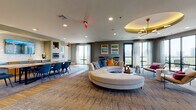 The Sky Lounge is a thoughtfully designed communal space that seamlessly blends elegance and functionality across a single, open floor. The main lounge area serves as the centerpiece, anchored by a large circular sectional sofa beneath a striking round ceiling medallion with a gold-toned coffered detail and a sculptural spur chandelier. Floor-to-ceiling windows wrap around this zone, flooding the space with natural light and offering expansive panoramic views of the surrounding cityscape and landscape.

Multiple distinct seating arrangements provide flexibility throughout the lounge. One area features contemporary accent chairs paired with a live-edge wood coffee table, rounded ottoman stools, and a wall-mounted flat-screen television set against marble-paneled surround. Decorative woven leaf wall art and a coffered teal ceiling treatment add an artistic, layered character to this corner. High-top bar seating along one wall, accompanied by curated colorful artwork, offers an additional casual gathering option.

The dining and kitchen zone is anchored by a striking live-edge communal table surrounded by teal upholstered chairs. Built-in appliances, including a wall oven, refrigerator, and wet bar with marble backsplash, make this area fully equipped for events and gatherings. Warm hardwood flooring in this section contrasts with the textured carpeting throughout the lounge, defining each zone while maintaining a cohesive, upscale aesthetic.
