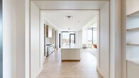 Located on S Second St in Minneapolis, MN, this elegantly designed single-floor space offers a seamless open-concept layout combining a kitchen and living area with refined, high-end finishes throughout. Herringbone-patterned hardwood flooring flows continuously across the entire space, anchoring the sophisticated design aesthetic.

The kitchen is a standout, featuring a lengthy waterfall-edge quartz island with integrated sink and brass fixtures, stainless steel professional-grade appliances, and a white subway tile backsplash. Flanking the range hood are dramatic built-in wine racks that extend nearly floor to ceiling, while a sculptural brass and white pendant light adds an artistic focal point overhead. Floor-to-ceiling windows with city views bring abundant natural light into the kitchen.

The living area is defined by a curved, channel-tufted leather sectional sofa in a muted lavender hue, accented by a sleek white rectangular coffee table. Built-in shelving frames a wall-mounted television, creating a polished media wall. An additional lounge zone features large windows draped in sheer white curtains, allowing soft, diffused light to fill the room. A dedicated dining area with a bold red table, warm-toned upholstered chairs, and cascading brass pendant lights completes this thoughtfully curated space, all set against sweeping views of the Minneapolis skyline.