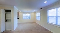 This single-floor residence offers a clean, classic aesthetic defined by thoughtful architectural details and a warm, welcoming ambiance throughout. The open living and dining area serves as the heart of the home, featuring soft neutral wall tones that complement the plush wall-to-wall carpeting underfoot. Crown molding adds a refined finishing touch along the ceiling perimeter, elevating the overall sense of quality and craftsmanship. Recessed lighting is strategically placed across the ceiling, providing even, adjustable illumination throughout the space, while a centrally hung chandelier introduces an elegant focal point to the dining area. Natural light fills the room generously through multiple windows fitted with white horizontal blinds, offering both brightness and privacy control. The windows frame views of the surrounding exterior, further enhancing the open, airy feel of the interior. A partial view of wood-toned cabinetry near the room's edge suggests a nearby kitchen, hinting at a practical and connected layout. White baseboards and trim work throughout tie the design together cohesively. Overall, this space presents a move-in ready environment with timeless design elements suited to a variety of furnishing styles.