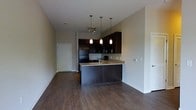 This single-floor space features an open-concept layout that seamlessly connects the kitchen and living area, creating a cohesive and functional environment. The rich, dark hardwood flooring runs throughout, providing a warm and polished foundation that contrasts elegantly with the neutral-toned walls.

The kitchen is a standout feature, anchored by a peninsula with granite countertops that doubles as a breakfast bar or prep space. Dark espresso cabinetry lines both the upper and lower sections of the kitchen, offering ample storage while contributing to the space's modern aesthetic. Black appliances, including a refrigerator and stove, complement the dark cabinetry. A set of pendant lights hangs above the peninsula, providing focused task lighting with a stylish touch. Additional pendant lighting clusters above the kitchen area further enhance illumination.

The open living area benefits from the kitchen's light spillover, creating a bright and airy atmosphere. A doorway on the left side of the space leads to additional rooms, while the overall layout is designed for efficient use of space without feeling cramped. The clean lines and contemporary finishes throughout give the space a cohesive, well-designed character.
