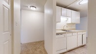 This single-floor residential unit offers a straightforward and functional layout designed for comfortable everyday living. The kitchen is positioned at one end of the unit and features white cabinetry extending to the upper walls, a built-in dishwasher, a stainless single-basin sink with a brushed nickel faucet, and under-cabinet lighting. Light-colored countertops and tile flooring complete the practical workspace.

The living area is open and generously sized, with warm beige carpeting underfoot and a textured ceiling overhead. A wall-mounted air conditioning unit provides climate control, while a baseboard heater runs along the perimeter wall. A sliding glass door or large window offers a view of greenery outside and connects to what appears to be a balcony or outdoor access point.

The bedroom areas are well-proportioned, each carpeted and finished with clean white walls and textured ceilings. Windows fitted with vertical blinds allow natural light to filter in, and baseboard heaters ensure year-round comfort.

The bathroom includes a wide vanity with a speckled countertop, white cabinetry below, an oval undermount sink, and a mirrored medicine cabinet with built-in shelving. Tile flooring ties the space together cleanly. Throughout, the unit maintains a neutral, light-toned palette that creates a bright and cohesive atmosphere.