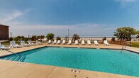 Located at 5617 Muirfield Dr SW in Cedar Rapids, Iowa, this outdoor pool area serves as a welcoming recreational amenity. The centerpiece is a generously sized swimming pool with clear turquoise water, marked at a depth of 5 feet and equipped with pool entry points for easy access. Surrounding the pool is a broad concrete sun deck that provides ample space for relaxation and movement. Numerous white lounge chairs and chaise lounges are arranged along all sides of the pool, offering plenty of seating for sunbathing and leisure. The open-air setting allows for abundant natural light and open sky views, contributing to a bright and airy atmosphere. The pool area is fully enclosed by a chain-link security fence, ensuring a safe and defined perimeter. A single-story clubhouse or amenity building with a pitched roof is situated directly adjacent to the pool, providing convenient access to additional facilities. The surrounding landscape includes neatly maintained green lawns and mature trees, with views of the multi-story residential buildings nearby. The overall design of the space prioritizes functionality and comfort, making it a practical and enjoyable outdoor gathering area for residents.