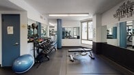 Located on W Newton St in Boston's South End neighborhood, this fitness center offers a comprehensive workout facility spread across a single floor. The space is designed with functionality and motivation in mind, featuring a clean, modern aesthetic with white walls accented by bold navy blue architectural detailing along the ceilings and trim.

The gym is organized into distinct workout zones. A dedicated free weights area features a full rack of dumbbells in a range of sizes, a stability ball, and flat benches, all set on durable rubber flooring. Large mirrors line the walls throughout, enhancing the sense of space and allowing users to monitor their form. Motivational fitness typography adorns the walls, adding to the energetic atmosphere.

A separate section houses a selection of cable and resistance machines suited for full-body strength training, arranged with ample space between equipment for comfortable use. Natural light filters in through multiple windows, providing views to the surrounding urban streetscape and keeping the environment bright and welcoming.

The entry area includes a body weight scale and informational postings, including heart rate target charts. Flush-mounted ceiling lights provide consistent illumination throughout. Access is managed through a key-entry blue door, contributing to the secure, controlled environment. The overall ambiance is clean, practical, and well-maintained, making it a capable fitness resource for its users.