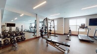 The fitness center at 40 Bennett Rd in Englewood, New Jersey, is a thoughtfully designed, single-floor facility offering a comprehensive range of exercise spaces and equipment. The layout is divided into distinct functional zones, each tailored to specific training needs.

The main strength training area features durable rubber flooring and is lined with large floor-to-ceiling mirrors that enhance the sense of space and allow users to monitor their form. This zone is well-stocked with a full rack of dumbbells in varying weights, a multi-function Smith machine, an adjustable weight bench, and additional free weight equipment. Linear LED lighting installed across the ceiling provides consistent, even illumination throughout.

An adjacent cardio area houses multiple treadmills and upright cardio machines positioned near windows that allow natural light to filter in. Wall-mounted flat-screen televisions are positioned for comfortable viewing during workouts.

A separate functional training studio features wood-look flooring, a wall-mounted TV, large mirrors, TRX suspension trainers, medicine balls, plyo boxes, and a dedicated equipment rack—ideal for group or circuit-style training. The studio benefits from recessed and pendant lighting, adding a polished aesthetic.

The facility is accessed through teal-framed glass double doors and includes a cable machine station and additional strength equipment near the entrance. The overall ambiance is clean, modern, and well-maintained.