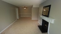 This single-floor space centers around a generously sized open living area finished with wall-to-wall carpet in a warm neutral tone. The room's most notable feature is a classic fireplace framed by a crisp white mantle and dark surround, accented by a large dark-framed mirror mounted above. The walls are painted in a soft taupe hue, complemented by clean white baseboards that run throughout the space, adding a subtle contrast. A ceiling fan with an integrated light fixture provides both air circulation and overhead lighting. The room extends into a secondary area, separated by a partial wall or step-down transition, which mirrors the same flooring and color palette, offering additional versatile living or dining space. A door visible in the background suggests access to an adjacent room or closet, adding to the functional layout. The overall ambiance is understated and neutral, providing a blank canvas well-suited for a variety of interior design styles. The consistent use of soft tones and carpet throughout creates a cohesive, calm atmosphere across the living space.