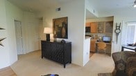 This single-floor residence offers an open-concept layout that seamlessly connects the main living area to the kitchen and a dedicated workspace nook. The living area features soft, neutral-toned carpet and smooth walls, providing a clean and versatile backdrop for furnishings. A bold, dark wood multi-drawer dresser serves as a focal accent piece beneath an abstract wall painting, adding character and contrast to the light interior palette. A closet with bifold doors is visible nearby, offering convenient built-in storage.

The kitchen is partially separated by a structural column, lending a sense of definition without fully closing off the space. It features warm honey-toned wood cabinetry, open shelving, light countertops, and standard appliances including a microwave. Adjacent to the kitchen is a compact built-in desk area with overhead cabinetry, ideal for use as a home office or study station. The desk nook accommodates a monitor, a chair, and additional shelving above, making it a highly functional addition to the layout. Overall, the space presents a practical and cohesive design with a warm, neutral ambiance throughout.