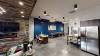 This thoughtfully designed amenity space delivers an impressive range of features across a single level, blending social, recreational, and professional environments seamlessly. The central lounge area makes an immediate impact with its striking reclaimed wood accent wall, built-in electric fireplace, and wall-mounted flat-screen TV, creating a warm and inviting gathering space. Bold cobalt blue walls punctuate the open recreation area, which includes a shuffleboard table, casual dining seating, and polished concrete floors that lend an industrial-modern aesthetic throughout. The fully equipped kitchen is a standout feature, showcasing dark cabinetry, stainless steel appliances including a large refrigerator and range, a herringbone tile backsplash, a spacious quartz island with bar seating, and elegant pendant lighting. A dedicated private screening room offers tiered leather recliner seating and acoustic wall panels, providing a true theater experience. For recreation enthusiasts, an indoor golf simulator room is outfitted with professional-grade equipment and a full projection screen. A formal conference room with a large boardroom table and wall-mounted display rounds out the space, making it equally suited for professional meetings. Additional casual seating nooks with high-top tables complete this versatile and well-appointed facility.