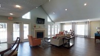 This single-story space is defined by its dramatic vaulted ceiling, which rises sharply to a peak and creates an expansive, airy atmosphere throughout the main living area. The ceiling is finished with a textured surface, and recessed lighting is strategically placed to illuminate the space effectively. Large transom windows flanking the peak of the vaulted ceiling allow generous natural light to flood the room, complementing the soft sage-green wall tones.

A central fireplace with a clean white mantel serves as the focal point of the living area, with a wall-mounted flat-screen television positioned above it. The dark hardwood flooring grounds the space and adds a warm contrast to the lighter walls and furnishings. The living area is arranged with a neutral sofa, a glass-top coffee table, and a pair of orange accent chairs, adding a vibrant pop of color to the otherwise understated palette.

French doors along the far wall introduce additional natural light and connect the interior to the outdoors. Adjacent to the living area, a dining space is furnished with dark wood chairs and a glass-top table, creating a seamless transition between the two functional zones. The overall ambiance is open, well-lit, and inviting.