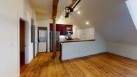 Located at 1329 Jefferson St in Lynchburg, Virginia, this single-floor unit offers a compelling blend of industrial heritage and contemporary living. Throughout the space, original exposed timber beams and historic pulley and belt-drive machinery have been preserved overhead, serving as striking architectural focal points that speak to the building's mill origins. Warm hardwood flooring runs consistently through the main living areas and hallway, complementing the raw character of the exposed elements.

The open kitchen features rich, deep-toned cabinetry, a butcher-block-style peninsula countertop, stainless steel appliances including a refrigerator, range, and over-the-range microwave, and recessed lighting above. A stacked washer and dryer unit is conveniently integrated into an adjacent alcove. The vaulted ceiling in this area adds a sense of vertical openness.

The living area is generously proportioned, with natural light filtering in through windows framed by the sloped roofline, exposed brick accents, and original structural iron columns. The hallway connects rooms efficiently while showcasing additional original pulley hardware overhead.

Bedrooms feature original beam detailing, hardwood floors, and closets with wire shelving. The bathroom offers tile flooring, a vanity with rich cabinetry matching the kitchen, and clean, neutral finishes throughout. This space delivers a distinctive, loft-inspired atmosphere rooted in Lynchburg's industrial history.