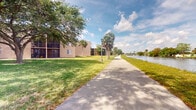 This compact space is part of a waterfront residential community characterized by its lush, well-maintained surroundings. The exterior environment features a paved pathway that runs alongside a calm body of water, flanked by tall palm trees and mature shade trees that provide natural canopy and visual appeal. The grounds are landscaped with manicured green grass, contributing to a serene, park-like atmosphere.

The multi-story buildings within the community are constructed with a neutral-toned brick or stucco exterior, featuring covered balconies that offer residents views of the surrounding landscape and waterway. Street-style lamp posts line the pathway, providing both functional lighting and a structured aesthetic to the outdoor corridor.

The interior images provided are too dark to detail specific features of the space itself. However, given the compact footprint, the space is best suited for focused, single-purpose use. Its single-floor layout ensures straightforward accessibility with no need for stairs or elevation changes.

Overall, the space benefits greatly from its setting within a well-kept, tropical-style community, where the natural environment and waterfront proximity contribute significantly to its ambient appeal.