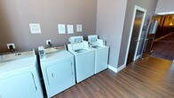 This single-floor shared laundry facility is designed with functionality and convenience in mind. The room features a lineup of top-loading washing machines paired with a full-size dryer, providing ample capacity for multiple users. The machines are neatly arranged along the wall, with posted informational notices displayed above them to guide users on proper usage and facility guidelines. The space is finished with warm hardwood-style flooring that adds a clean, polished look to an otherwise utilitarian room. Neutral-toned walls in a muted taupe shade contribute to a tidy and well-maintained atmosphere. White baseboards provide a crisp contrast to the flooring, reinforcing the clean aesthetic throughout. The layout is efficient, allowing easy access to each machine, and the overall condition of the room reflects careful upkeep. This shared laundry room offers a straightforward, no-frills experience well-suited for residents or guests seeking convenient on-site laundry access.