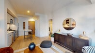 This well-appointed one-bedroom residence combines contemporary design with functional living across a single floor. The open-concept living area features light hardwood flooring, crisp white walls, and a cohesive layout that connects the living space to the kitchen and entryway. A bar-height dining table with stools sits adjacent to the kitchen, offering a casual dining option, while the living area is anchored by a woven area rug, a dark wood credenza, and a round mirror that adds depth to the space.

The kitchen is efficiently designed with light grey cabinetry, white quartz countertops, and a geometric elongated hexagon tile backsplash. Open wooden shelving, stainless steel appliances—including a gas range, built-in microwave, dishwasher, and refrigerator—complete this well-equipped culinary space.

The bedroom is a serene retreat, highlighted by expansive floor-to-ceiling windows that frame urban city views and flood the room with natural light. The bathroom showcases striking dark hexagon mosaic floor tiles, a floating vanity with wood-trimmed cabinetry, a large framed mirror, and a shower-tub combination with white subway tile surround—offering a clean, modern aesthetic throughout.