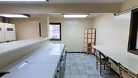 This shared laundry facility is designed to provide a practical and comfortable environment for everyday use. The room is finished with light yellow walls and neutral tile flooring, creating a clean and utilitarian atmosphere. Overhead fluorescent panel lighting ensures the space is well-lit throughout.

A row of full-sized top-loading washers and front-loading dryers lines one wall, offering ample capacity for laundry needs. A long, flat folding surface runs adjacent to the machines, providing convenient space for sorting and folding clothes. A small window along the same wall allows natural light to filter in and offers a view of the surrounding exterior.

On the opposite side of the room, a wooden tri-fold room divider serves as an organizational station, appearing to hold informational sheets or notices. Nearby, a table paired with wooden chairs provides a comfortable waiting area. The overall layout is thoughtfully arranged to accommodate multiple users simultaneously, balancing functional equipment with practical amenities in a straightforward, well-maintained setting.