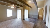 This single-floor space makes a strong architectural impression with its vaulted ceilings adorned by prominent exposed wood beams, lending a rustic yet refined character to the interior. The open living area is generously proportioned, seamlessly transitioning between plush carpeting and rich dark hardwood flooring, offering both comfort and visual contrast.

Natural light flows abundantly into the space through multiple large windows and two sets of sliding glass patio doors fitted with vertical blinds, brightening the neutral-toned walls that feature a two-tone paint scheme — a warm beige above and a deeper accent tone below a dividing line. A ceiling fan with a light fixture is centrally positioned, adding both function and a casual charm to the room.

Track lighting along the ceiling provides additional illumination, complementing the natural light. The dual sliding glass doors suggest direct outdoor access from the main living area, enhancing the sense of openness and connectivity to the exterior. The combination of architectural details — from the dramatic beamed ceiling to the contrast of flooring materials — creates a warm and inviting atmosphere well-suited for everyday living.