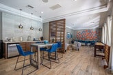 Located at 13120 Day St in Moreno Valley, CA, this single-floor community amenity space is thoughtfully designed to encourage socializing and relaxation. The open-concept layout is anchored by wide-plank hardwood-style flooring and elegant crown molding throughout, creating a warm yet polished atmosphere.

A kitchenette area features warm wood cabinetry, a concrete-top bar table with vibrant blue rope barstools, pendant lighting in brushed silver, subway tile backsplash, and stainless steel appliances — ideal for casual gatherings. A slatted wood partition screen serves as a stylish architectural divider between the kitchenette and adjacent lounge zones.

The dining area is centered around a substantial wooden table surrounded by tan leather chairs, complemented by plush teal accent chairs, a bold modern chandelier, and curated wall art in large frames. A separate lounge nook provides a cozy retreat with a wall-mounted TV, media console, layered seating, and vibrant decorative accents in teal and yellow.

The games area runs along a wall of floor-to-ceiling windows and glass doors that flood the space with natural light and offer direct views of the outdoor pool and courtyard. A shuffleboard table and oversized connect-four game add a playful, social dimension. Sheer linen drapes frame the windows, enhancing the airy, resort-inspired ambiance of the entire space.