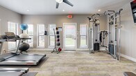 This dedicated fitness center offers a thoughtfully designed workout space with a neutral gray-toned color palette and modern finishes throughout. The room features durable, patterned carpet tiles in gray and yellow tones, recessed lighting, and a ceiling fan to ensure a comfortable environment during exercise.

The space is comprehensively outfitted with a wide variety of equipment to support diverse fitness routines. Cardio options include a treadmill, elliptical trainer, and a rowing machine, while strength training is well-represented with a cable machine, a multi-tier dumbbell rack, a kettlebell rack, and an adjustable workout bench. A medicine ball storage rack adds additional versatility for functional training. Wall-mounted yoga or exercise mats provide a convenient stretching and floor exercise option.

A wall-mounted flat-screen television enhances the workout experience, while a security camera ensures a safe environment. Multiple windows fitted with white shutters line one wall, allowing ample natural light and outdoor views of the surrounding greenery. A dedicated exterior door provides direct access to the outdoors. French doors connect the fitness center to an adjacent lounge or common area, creating a seamless flow between spaces. The overall ambiance is clean, bright, and purposefully designed to motivate and support an active lifestyle.