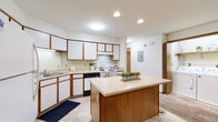 This single-floor unit offers a practical and comfortable layout with a range of functional spaces throughout. The kitchen is a standout area, featuring white cabinetry with warm wood trim accents, light countertops, tile flooring, recessed lighting, and a central island that provides additional prep and dining space. White appliances, including an electric range and dishwasher, are neatly integrated into the layout, and an in-unit washer and dryer are conveniently located just off the kitchen. The living area is finished with wall-to-wall carpet and neutral walls, creating a clean, open atmosphere. A glass-paned exterior door brings in natural light and adds character to the space. The bedrooms are carpeted and generously sized, with one featuring a large picture window overlooking a brick exterior and a reach-in closet, providing ample storage. Two bathrooms serve the unit—one featuring a vanity with cabinet storage and tile flooring, and the other a walk-in shower stall with a wall-mounted mirror. A carpeted staircase with a warm wood handrail connects to additional levels. Throughout, warm wood trim and door accents provide a cohesive, classic aesthetic.