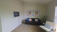 This single-floor space offers a clean, modern aesthetic anchored by a welcoming living area. The room features neutral, warm-toned walls and plush carpeted flooring that contribute to a calm and inviting ambiance. A dark upholstered sofa serves as the centerpiece, accented with vibrant blue and floral throw pillows that add a pop of color against the understated backdrop. A wall-mounted flat-screen television is positioned for comfortable viewing, while two colorful framed art pieces displaying bold geometric designs hang prominently on the wall, adding personality and visual interest to the space. A sleek white coffee table and a decorative branched arrangement in the corner complete the living area's refined yet approachable style. The room benefits from generous wall height, giving it an open, airy feel. The overall design balances simplicity with carefully chosen decorative touches, creating a space that feels both functional and thoughtfully curated. The neutral color palette and consistent finishes throughout suggest a cohesive interior design approach that would suit a variety of personal styles.