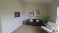 This single-floor space offers a clean, modern aesthetic anchored by a welcoming living area. The room features neutral, warm-toned walls and plush carpeted flooring that contribute to a calm and inviting ambiance. A dark upholstered sofa serves as the centerpiece, accented with vibrant blue and floral throw pillows that add a pop of color against the understated backdrop. A wall-mounted flat-screen television is positioned for comfortable viewing, while two colorful framed art pieces displaying bold geometric designs hang prominently on the wall, adding personality and visual interest to the space. A sleek white coffee table and a decorative branched arrangement in the corner complete the living area's refined yet approachable style. The room benefits from generous wall height, giving it an open, airy feel. The overall design balances simplicity with carefully chosen decorative touches, creating a space that feels both functional and thoughtfully curated. The neutral color palette and consistent finishes throughout suggest a cohesive interior design approach that would suit a variety of personal styles.