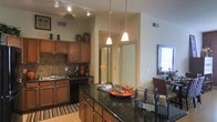 This single-floor space centers around a well-appointed kitchen that combines functionality with an inviting aesthetic. The kitchen features rich, warm-toned wood cabinetry that extends to the upper walls, providing generous storage throughout. A classic stone tile backsplash adds texture and visual interest behind the cooking area, complementing the dark, polished granite countertops that define the extended kitchen island. The island serves as both a prep surface and a casual dining or gathering area, with seating positioned along one side.

Overhead, a combination of recessed track lighting and elegant pendant lights hang at varying heights above the island, casting a warm, layered glow across the space. The flooring transitions between a striped area rug near the main cooking zone and light hardwood extending toward adjacent areas. A hallway visible beyond the kitchen suggests a thoughtful open layout connecting to additional rooms. Decorative accents, including ceramic vessels displayed atop the upper cabinets, add a personal, curated touch. The overall ambiance is warm and functional, with a cohesive design palette of earth tones and dark accents throughout.