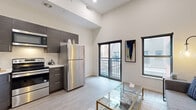 This thoughtfully designed one-bedroom, one-bathroom unit maximizes its compact footprint through an efficient open-concept layout and notable architectural details. The combined kitchen and living area forms the heart of the unit, distinguished by dramatically high ceilings that amplify the sense of space well beyond its physical dimensions. Light-toned hardwood-style flooring runs throughout, complementing the neutral white walls.

The kitchen is outfitted with a full suite of stainless steel appliances, including a refrigerator, electric range, built-in microwave, and dishwasher. Dark wood-grain cabinetry extending to the ceiling provides ample storage. The living area benefits from a sliding glass door that opens to a private balcony, alongside an additional window, flooding the space with natural light and outdoor views.

The bedroom is a cozy, well-defined space with a high clerestory window for light and privacy. The bathroom features large-format tile flooring, a frameless sliding glass shower enclosure with a built-in bench, a wall-mounted vanity with an undermount sink, and a wide mirror, delivering a clean, contemporary aesthetic throughout.
