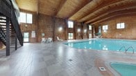 This single-floor indoor aquatic facility at Woodmere offers a generously sized swimming pool set within a striking rustic architectural environment. The space is defined by its soaring wood-beamed ceilings and warm, wood-paneled walls that extend from floor to ceiling, creating a cabin-like yet refined atmosphere. The pool itself features clear, well-maintained water with clearly marked depth indicators, surrounded by a neutral-toned tile deck that provides ample circulation space on all sides. Natural light filters into the space through a series of windows positioned at both standard and clerestory heights along the walls, complementing the wall-mounted lighting fixtures throughout. The lounge area is furnished with white slatted chairs, chaise lounges, and patio tables topped with branded green Woodmere umbrellas, creating comfortable spaces for relaxation. A metal staircase leads to an elevated mezzanine-level observation or utility area, adding vertical interest to the space. The facility also includes a smaller secondary pool or hot tub area visible at one end, along with essential amenities such as a waste receptacle and posted safety signage. Windows along the lower walls offer views of the surrounding greenery outside, reinforcing the connection to the natural landscape.